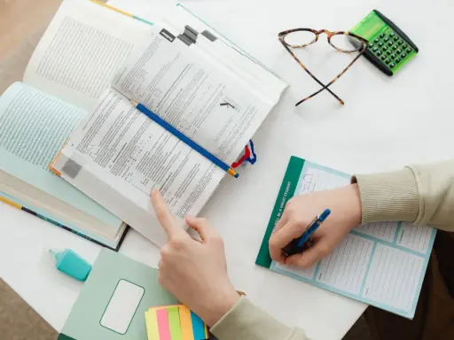 Uczeń pochyla się nad książkami, przygotowując się do egzaminu licencjackiego lub magisterskiego. Na stole leżą notatki, okulary i kalkulator.