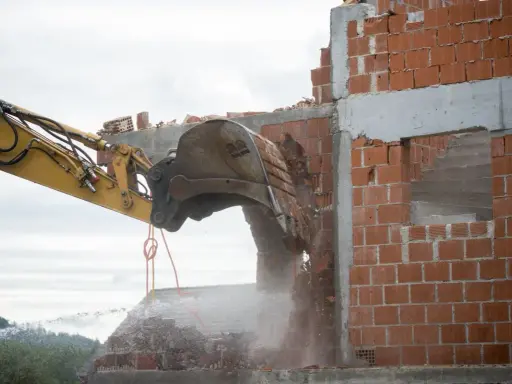 Koparka niszczy ceglaną ścianę. Rozbiórka postępuje, a gruz spada w dół.