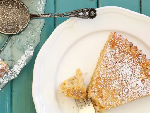 Saftige spanische Mandeltorte, bestäubt mit Puderzucker, auf einem weißen Teller. Ein Stück wurde bereits mit einer Gabel entnommen.