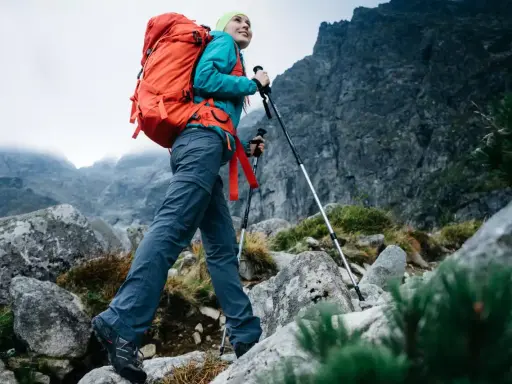 Kobieta z plecakiem wyrusza na wypad w góry, wspierając się kijkami trekkingowymi po kamienistym szlaku.