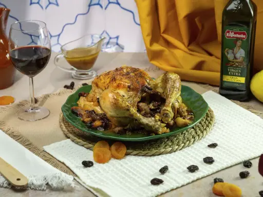 Pollo asado relleno, dorado y jugoso, servido en plato verde con pasas y orejones. Acompañado de vino tinto y aceite de oliva.