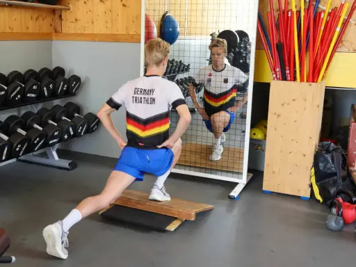 Ein Athlet im "Germany Triathlon"-Shirt macht Ausfallschritte im Athletik Training. Hanteln und Trainingsgeräte im Hintergrund.