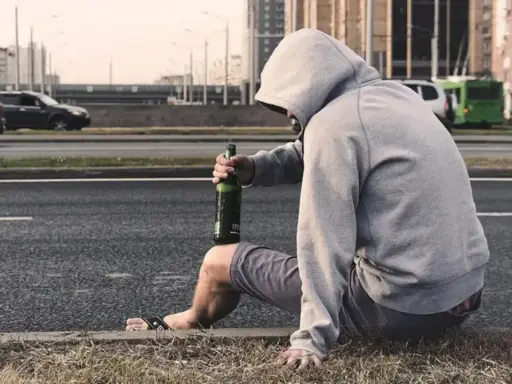 Mężczyzna w kapturze siedzi przy drodze z butelką piwa. Jego postawa sugeruje walkę z nałogiem, być może potrzebuje pomocy terapeuty od uzależnień.