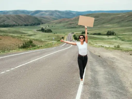 Kobieta z kartonem i kciukiem w górze na pustej drodze. Uczy się, jak łapać stopa, podróżując przez malownicze krajobrazy.