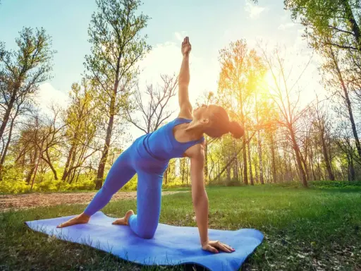 Kvinna utför yoga utomhus i en skogsmiljö. Solen skiner genom träden.
