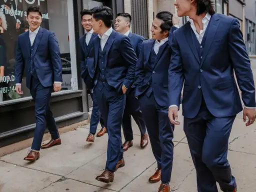 A group of men in navy suits walk down a sidewalk. They are all wearing brown dress shoes, proving you can wear brown shoes with a navy suit.