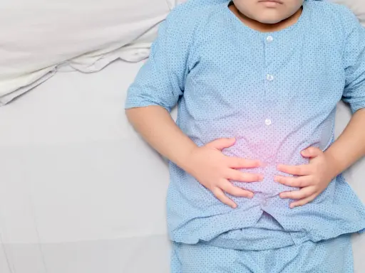 Niño con pijama azul, acostado en cama blanca, se agarra la barriga con las manos. Se visualiza un área rosada en su abdomen, indicando dolor barriga niños.