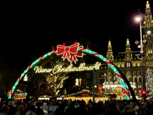 Wiedeński jarmark świąteczny w blasku świateł. Tłumy ludzi spacerują pod łukiem z napisem "Wiener Christkindlmarkt", w tle widać zabytkowe budynki i choinkę.