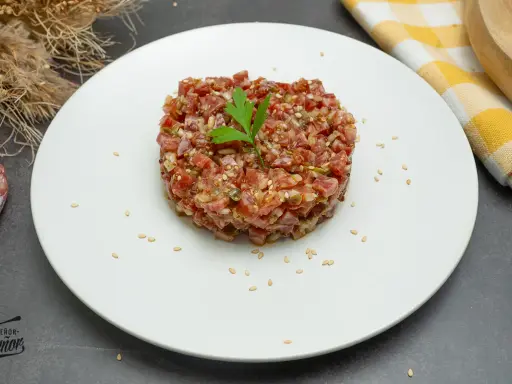 Tartar de fuet picado finamente, con semillas de sésamo y perejil, servido en plato blanco.