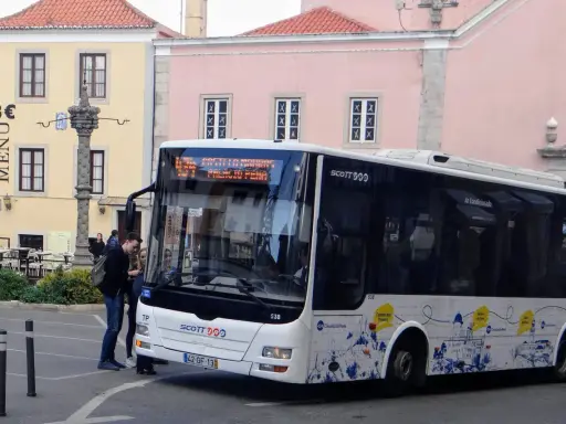 Ein Scott-Bus in Portugal hält an einer Haltestelle. Touristen steigen ein, während im Hintergrund bunte Gebäude und ein historischer Brunnen zu sehen sind.