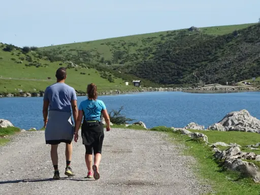 Ein Paar wandert auf einem Schotterweg am Ufer eines Sees. Grüne Hügel und Felsen prägen die **Spanien Natur**.