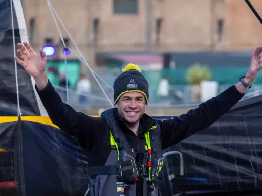 Nicolas d'Estais, marin souriant, bras levés, porte un bonnet et un gilet de sauvetage, prêt pour l'aventure en mer.