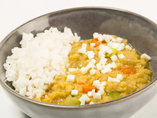 Un tazón de arroz blanco junto a un guiso cremoso de lentejas rojas con trozos de queso y verduras.