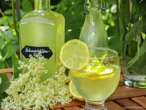 Erfrischendes Glas mit selbstgemachtem Holunderblütensirup, Zitrone und Minze. Daneben Flaschen mit dem Sirup und frische Holunderblüten.