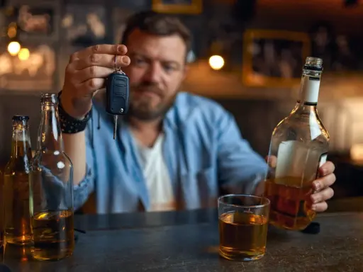Mężczyzna trzyma kluczyki do samochodu, zastanawiając się, czy jestem pijany. Obok butelki i szklanka alkoholu.
