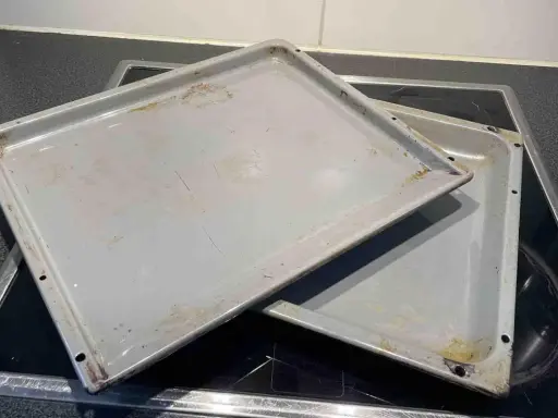 Zwei eingebrannte Backbleche liegen auf einem Cerankochfeld. Zeit, sie zu reinigen und wieder glänzen zu lassen!