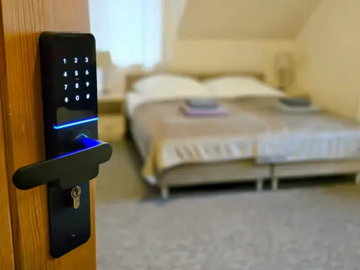 Cyfrowa klamka z klawiaturą numeryczną i niebieskim podświetleniem, symbolizująca digitalizację w hotelarstwie. W tle łóżko.