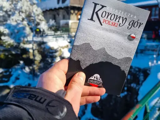 Dłoń trzyma książeczkę "Korona Gór Polski" na tle zimowego krajobrazu.