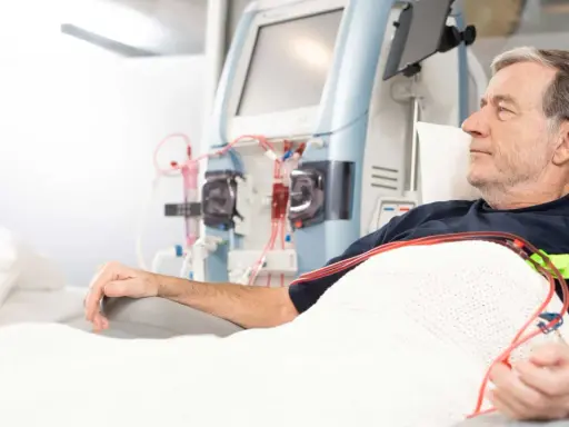 Starszy mężczyzna leży pod kocem, podłączony do aparatury do dializy. Jego ręka spoczywa na podłokietniku, a obok widać maszyny i czerwone wężyki.