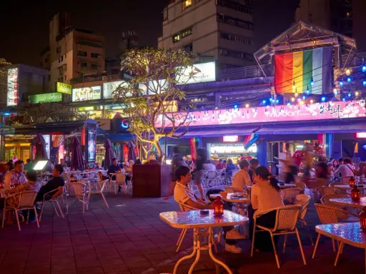 A vibrant night scene in gay Taipei, with people dining outdoors under colorful lights and a rainbow flag.
