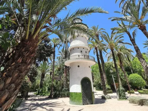 Torre blanca con balcón y puerta verde, rodeada de palmeras y vegetación exuberante. Un lugar encantador que ver en Elche.