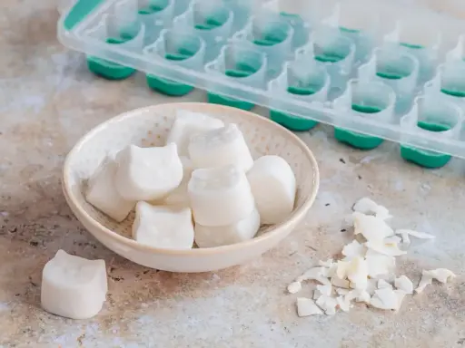 Kleine, weiße Eiswürfel in Katzenform liegen in einer Schale. Sie sind das Ergebnis von kokosmilch einfrieren. Daneben eine leere Eiswürfelform.