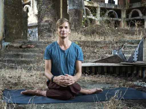 Man i blå tröja sitter i lotusställning på en yogamatta i ruiner. En stunds natthiko meditation i en övergiven miljö.