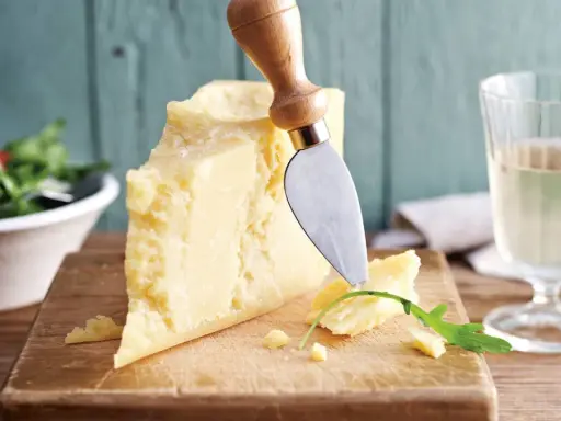 Ein Stück Parmesan, perfekt für vegetarische Gerichte, wird mit einem Käsemesser angeschnitten. Daneben ein Glas Weißwein und ein Salat.