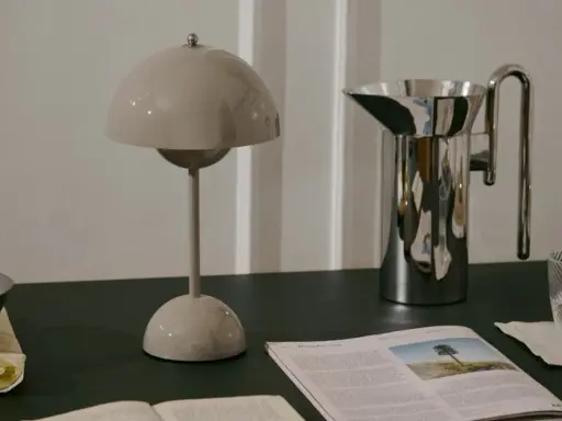 En beige lampa i studio lawahl-stil står bredvid en blank vattenkanna. En tidning med ett träd på omslaget ligger framför.