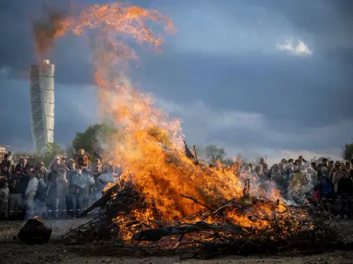 Stort bål med flammor som slår upp mot en dramatisk himmel. En folkmassa samlas för att bevittna ett traditionellt valborg firande.