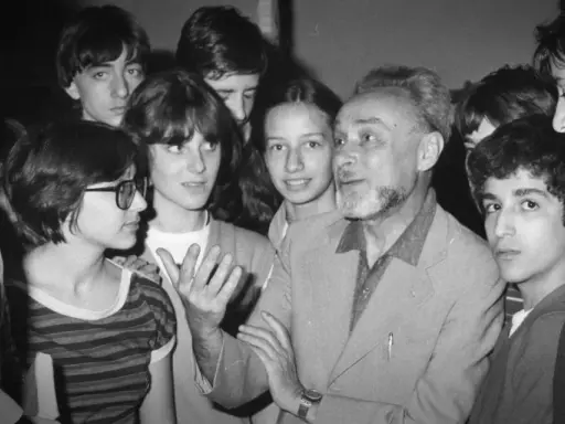 Un uomo anziano con barba e capelli grigi parla animatamente a un gruppo di giovani. Se questo è un uomo riassunto, è un uomo che ispira e coinvolge.