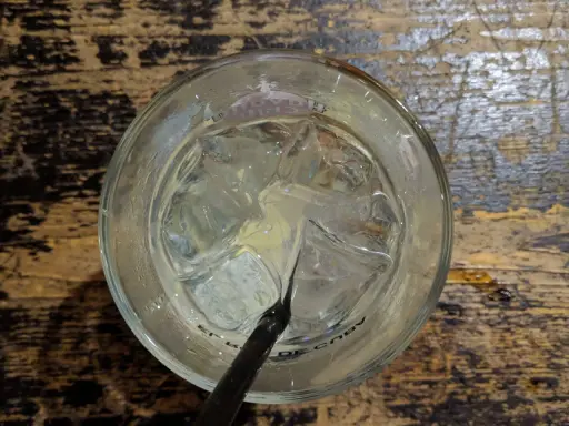 Ein Glas mit Eiswürfeln und einem schwarzen Strohhalm, bereit für einen erfrischenden Gin Tonic auf rustikalem Holztisch.