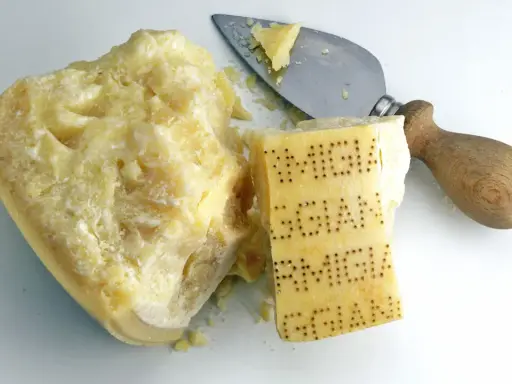 Ein Stück Parmesan mit eingeprägtem Schriftzug und ein Käsemesser. Aus was besteht Parmesan? Aus Milch, Lab und Salz.
