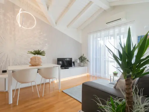 Luminoso appartamento mansardato con travi a vista, zona giorno con tavolo, sedie, TV e divano. Parete con carta da parati decorativa.