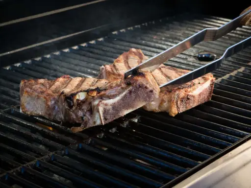 Zwei saftige Cowboy-Steaks werden mit einer Zange auf einem heißen Grill gewendet. Rauch steigt auf, während die Flammen darunter lodern.