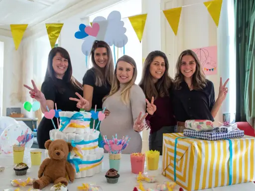 Pięć uśmiechniętych kobiet świętuje baby shower. Na stole tort z pieluch, prezenty i słodkości. To radosne wydarzenie, co to jest baby shower, pełne miłości i oczekiwania.