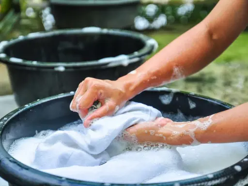 Manos lavando ropa blanca a mano en una tina con espuma, un método tradicional que explica qué significa lavado a mano en lavadora.