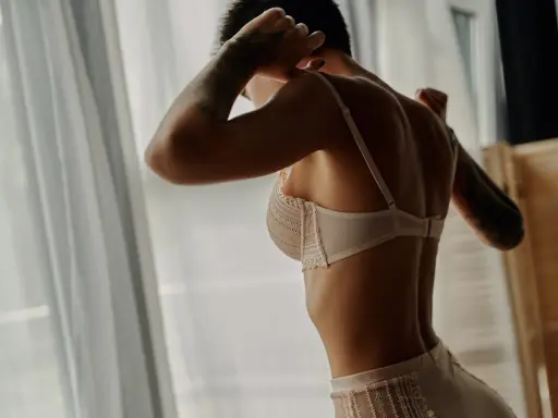 Donna con tatuaggi, indossa un reggiseno pizzo che esalta la scollatura seno. Luce soffusa da finestra.
