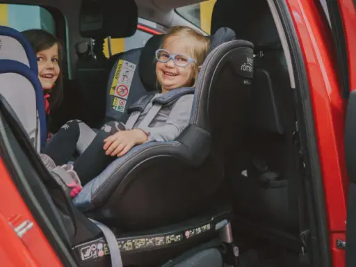 Dwie uśmiechnięte dziewczynki w fotelikach Britax Romer 0-18 kg w czerwonym samochodzie.