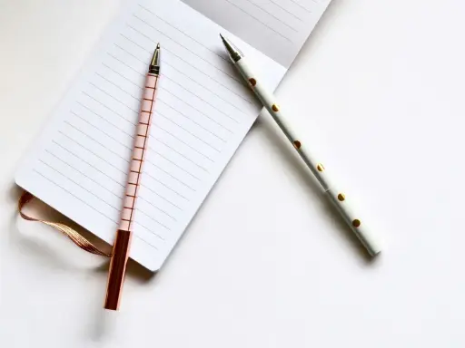 Deux stylos élégants, un rose à carreaux et un blanc à pois dorés, reposent sur un carnet ligné, prêts pour les éditions Balland manuscrit.