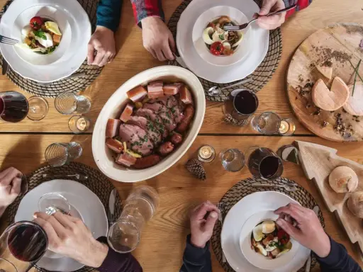 Un repas du terroir avec viande, saucisses, fromages et pain sur une table en bois. Des mains servent et dégustent.