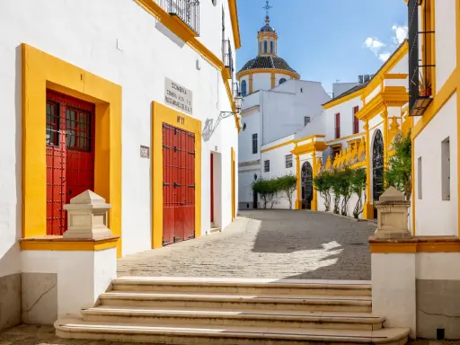 En solig gränd i Sevilla, Spanien, med vita hus, gula detaljer och röda dörrar. En kupol syns i bakgrunden.