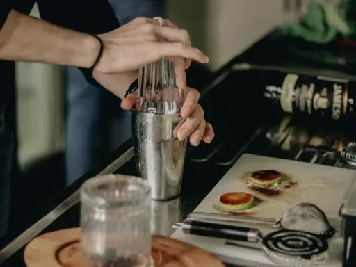 Ręce mieszają składniki w shakerze, przygotowując drinki na ciepło. Na blacie leży butelka whiskey i przypalone plasterki jabłka.