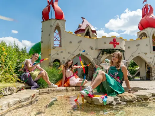 Trzy wróżki w kolorowych strojach siedzą przy strumyku przed bajkowym zamkiem. To z pewnością jedne z wielu magiczne ogrody atrakcje.