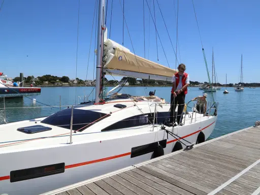 Un homme en gilet rouge ajuste une aussière sur un bateau blanc, prêt à appareiller. D'autres voiliers sont au mouillage.