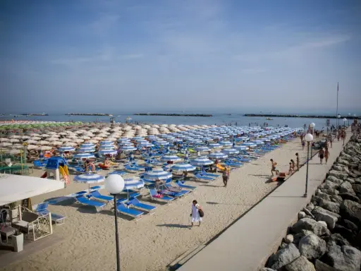 Spiaggia affollata a Rimini Gatteo Mare, con file ordinate di ombrelloni e lettini blu e bianchi, bagnanti e un molo di roccia.