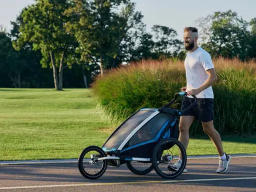 Ein Mann joggt mit Kinderwagen durch eine grüne Parklandschaft.