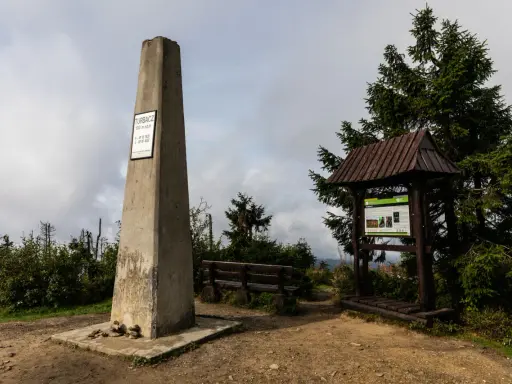 Kamienny obelisk na szczycie Turbacz w Gorcach, obok drewniana wiata z tablicą informacyjną i ławką.