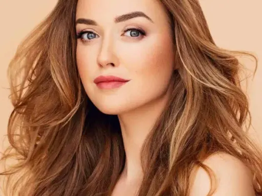 Mujer con cabello castaño dorado, ondulado y con reflejos claros. Su mirada es penetrante y sus labios pintados de rosa.