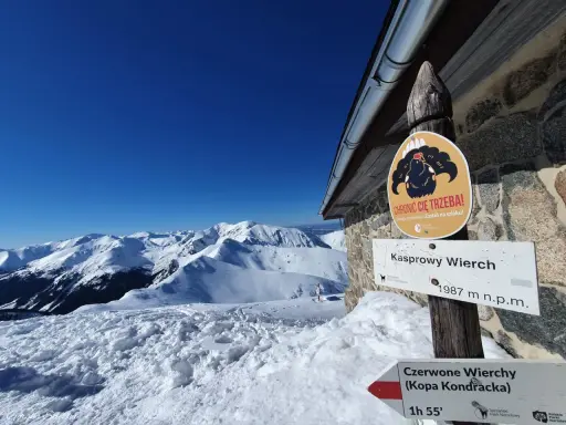 Zimowy krajobraz na Kasprowym Wierchu. Znaki kierunkowe wskazują drogę, a tablica przypomina o ochronie cietrzewia.